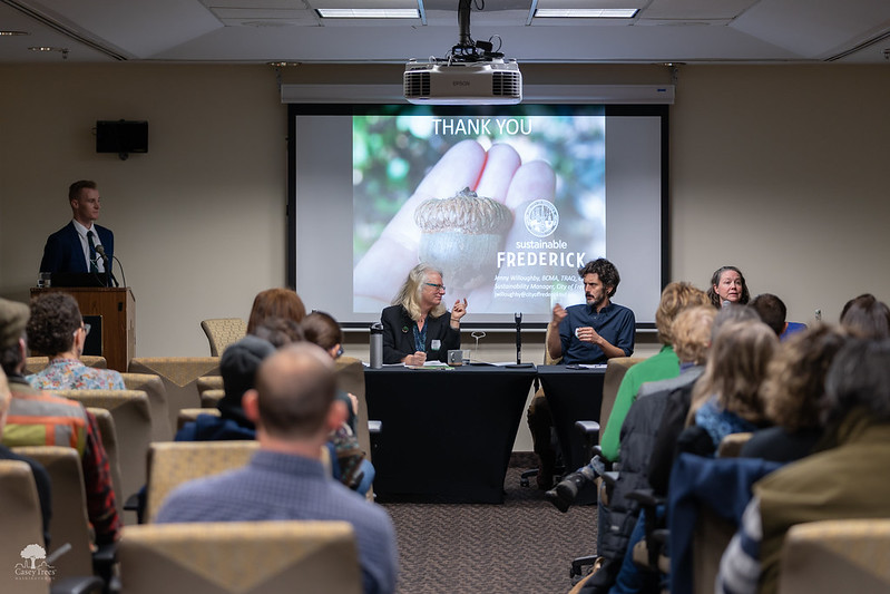 A discussion with 2 people at a table, someone at a podium, and an audience in a classroom