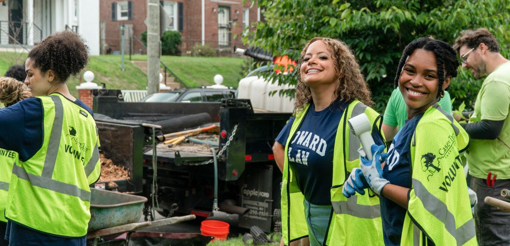 Two incoming Howard University Law students smile for the camera in front of a truck filled with mulch. Other students and Casey Trees employees work in the background.
