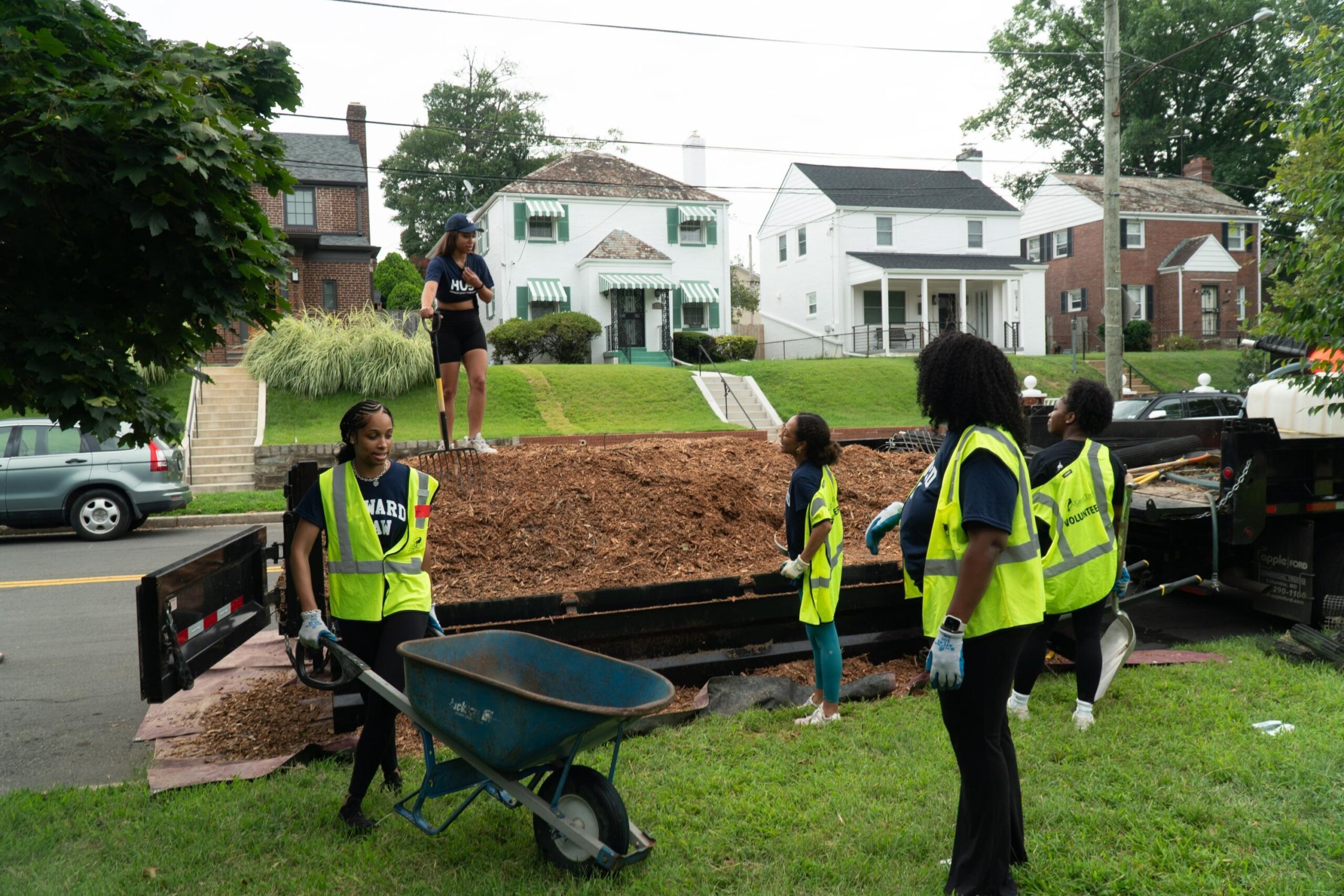 Five Howard University Law students work on moving mulch from the truck. One student is moving a wheelbarrow. Another student is standing on top of the mulch pile, holding a pitch fork, and giving instructions to three students who stand and listen.
