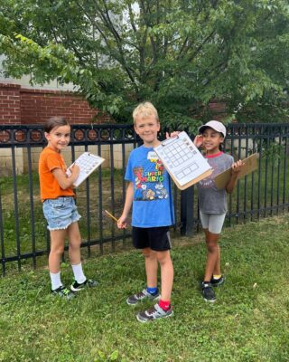 Three young kids smile for the camera and hold up their clipboards as they play a nature identification game outside.