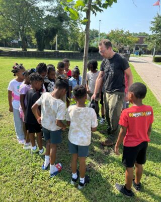 A group of students watch as a member of Casey Trees staff waters a young tree with a watering can.