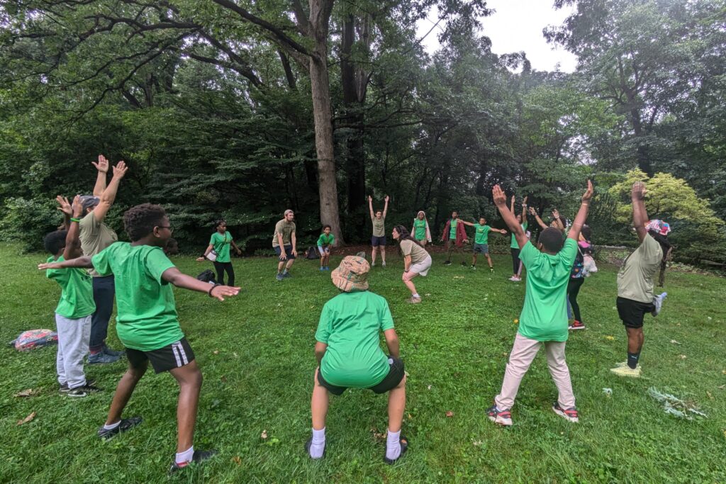 A group of students and educators stand in a circle outside as they play a nature game.