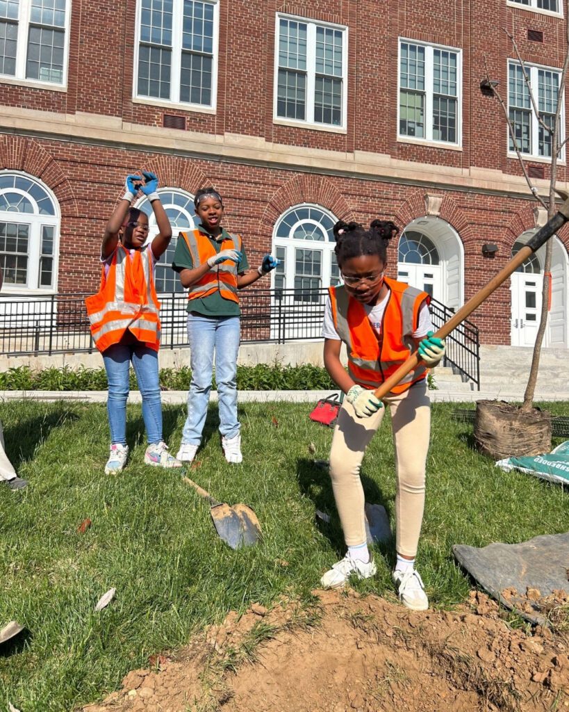 A student pickaxes the ground as two students cheer on.